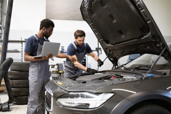 Mechanics in workshop repairing vehicle engine using laptop for diagnostics. Teamwork and technology in automotive repair service. Modern car maintenance and inspection.